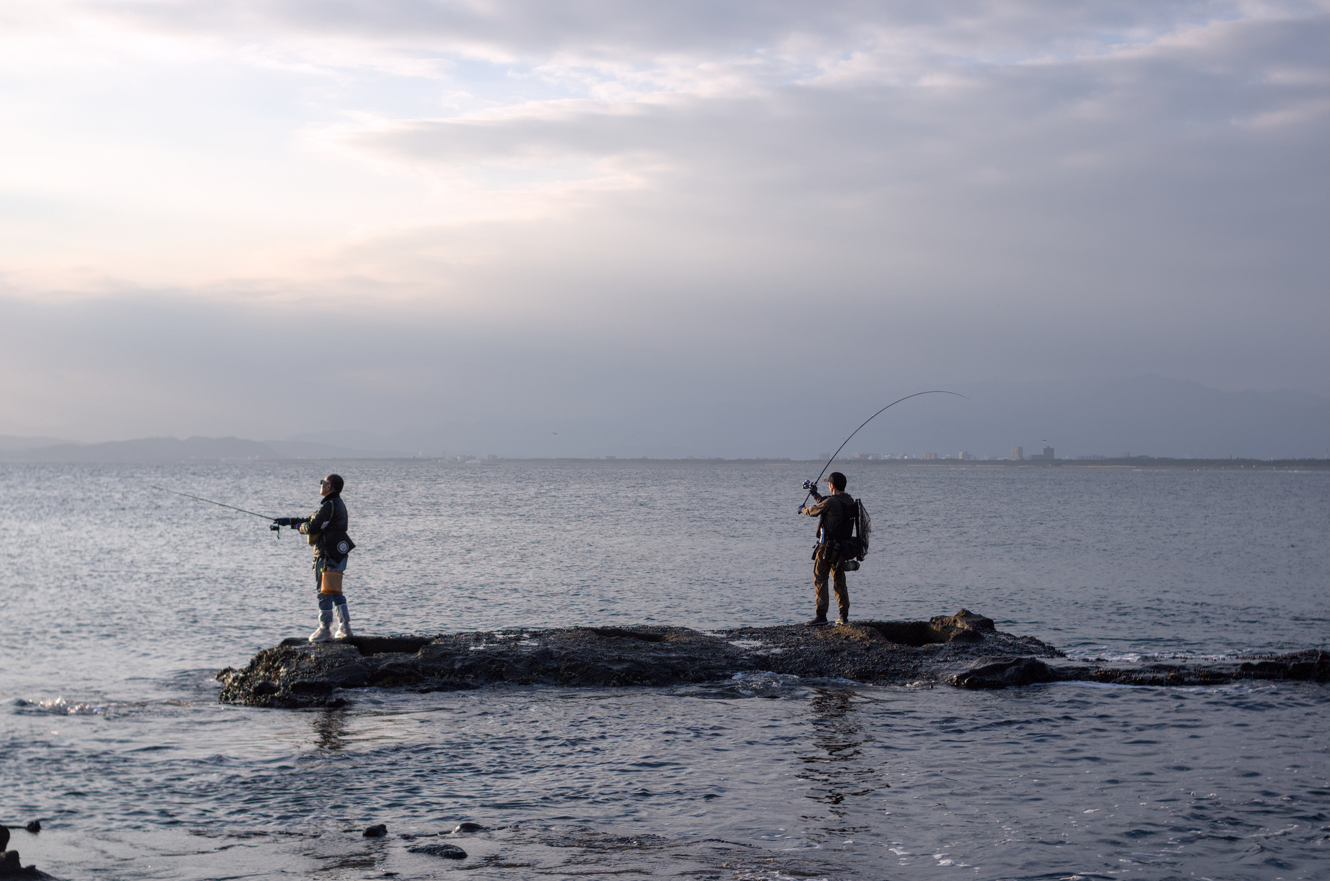 Two fishermen on a rock, one whipping their fishing rod forward.